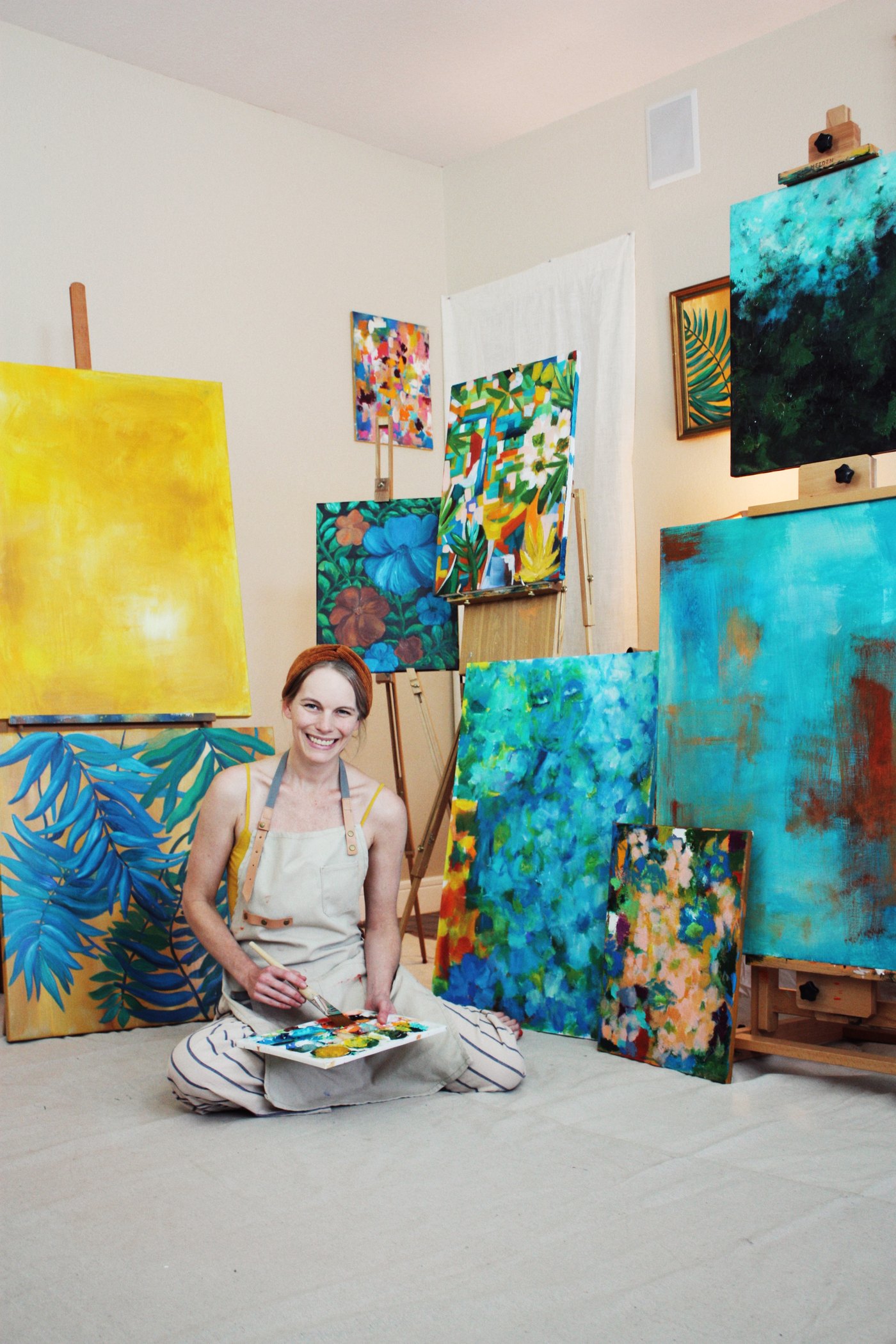 Kate Yarter sitting on the studio floor with a palette in her lap, her paintings stacked and leaning around her