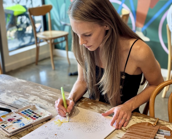 Kate Yarter painting a watercolor at a wooden table, pan palette open beside her